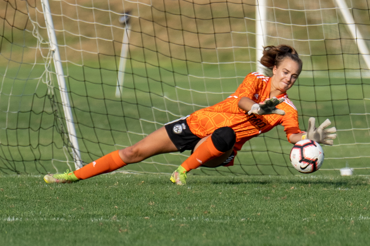 Female goalkeeper wearing an orange jersey, black shorts and orange socks, diving to save a soccer ball during a game.